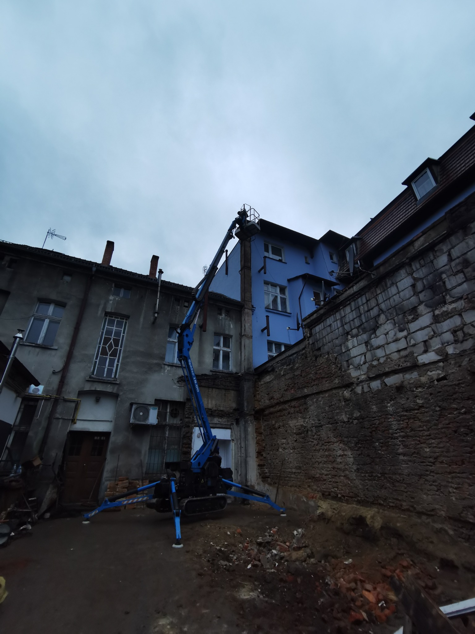 Niebieski podnośnik koszowy Spider 18.90 ustawiony na nierównym terenie między budynkami, z platformą roboczą na wysokości elewacji niebieskiego budynku.