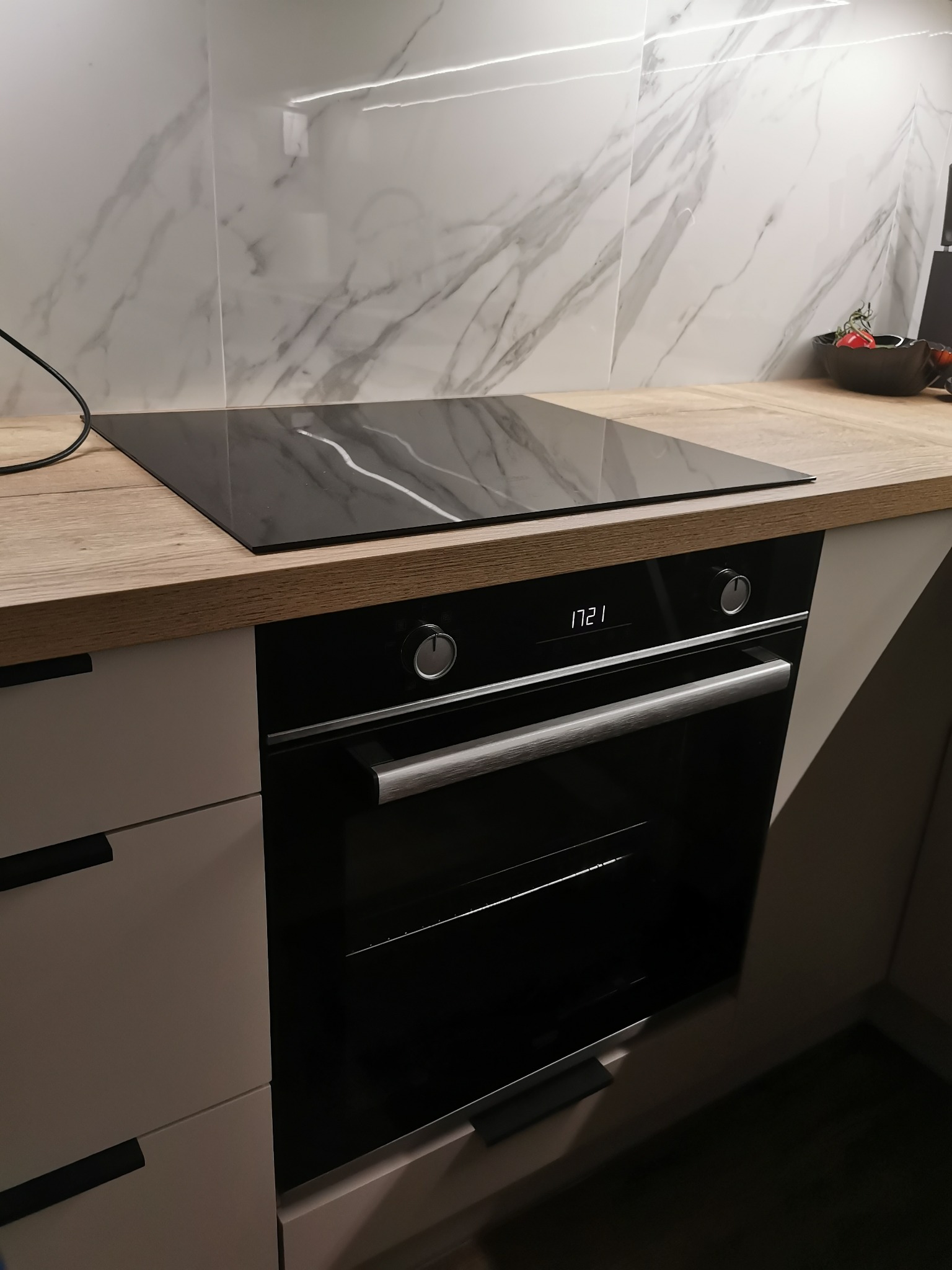 Nowoczesna kuchnia z wbudowaną płytą indukcyjną i piekarnikiem, białe marmurowe płytki na ścianie, drewniany blat, czarne uchwyty szafek.