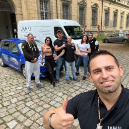 JAMAL Sp. z o.o. - Grupa pracowników firmy budowlanej Jamal pozuje do zdjęcia typu selfie na tle samochodów firmowych i budynku z cegły, jeden z mężczyzn pokazuje kciuk w górę.