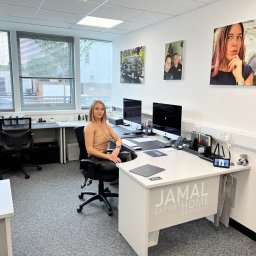 JAMAL Sp. z o.o. - Wnętrze biura z blondynką siedzącą przy biurku z dwoma monitorami, na ścianach wiszą obrazy z logiem firmy Jamal Expert Home Services