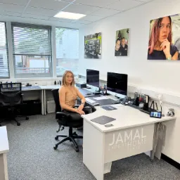 Wnętrze biura z blondynką siedzącą przy biurku z dwoma monitorami, na ścianach wiszą obrazy z logiem firmy Jamal Expert Home Services