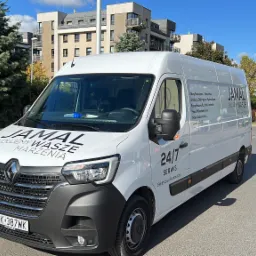 Biały samochód dostawczy firmy Jamal z napisem 'Budujemy Wasze Marzenia' i numerem telefonu, zaparkowany na ulicy w słoneczny dzień, na tle budynków mieszkalnych i drzew.