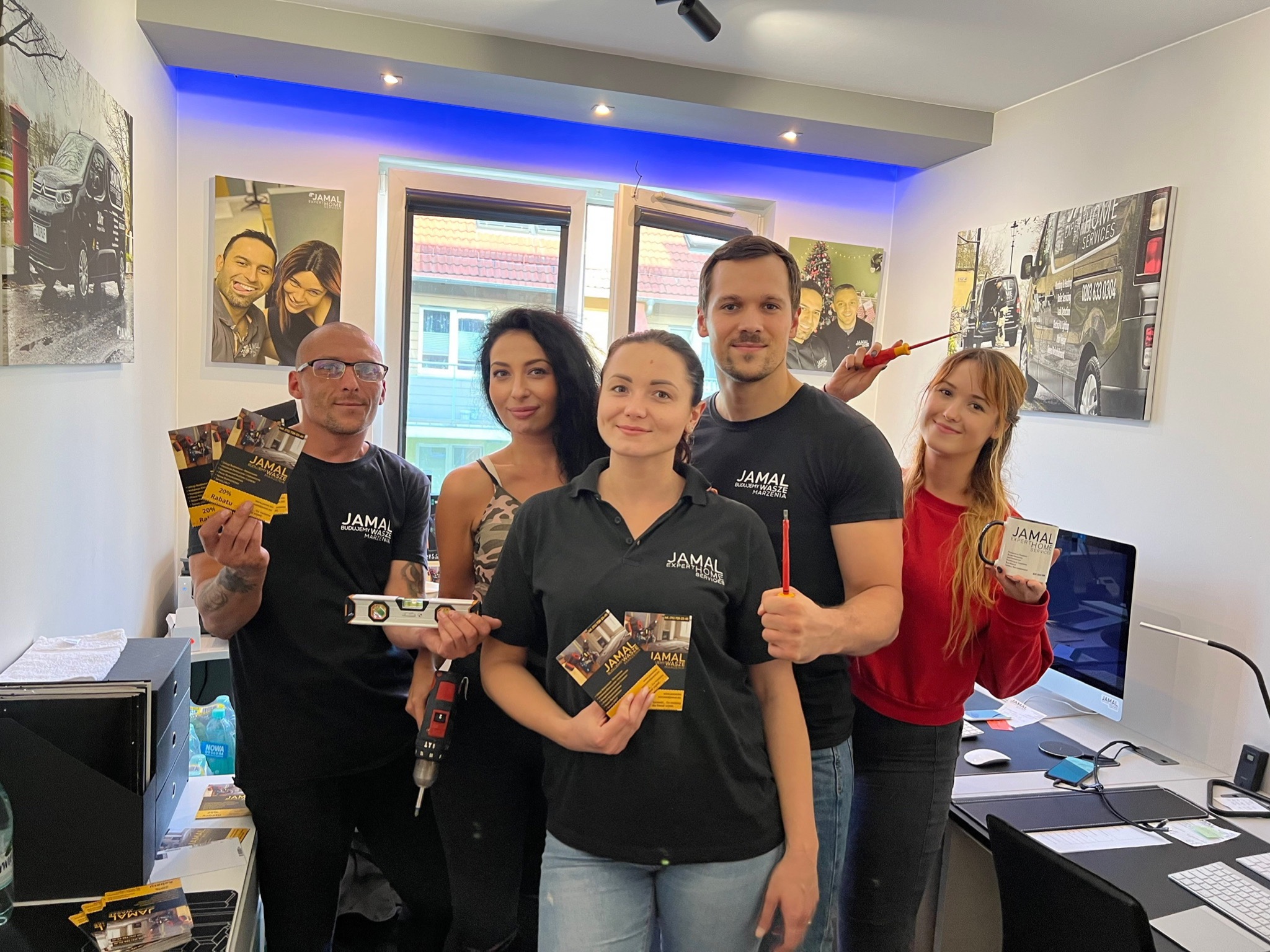Grupa pięciu osób z firmy Jamal Expert Home Services pozuje w biurze, trzymając wizytówki, poziomice, wkrętarkę i kubek z logo firmy, na ścianach wiszą obrazy z logo i pojazdami firmy.