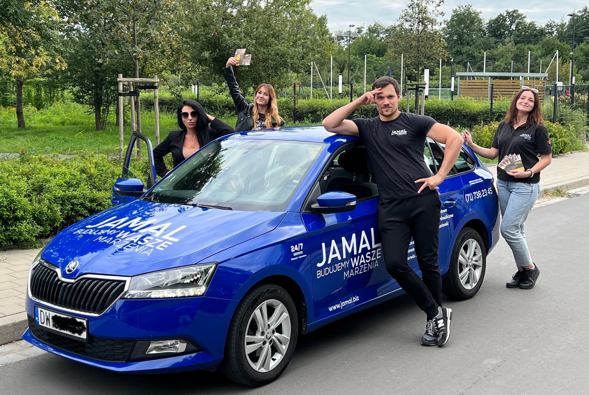 Zespół firmy budowlanej JAMAL prezentuje się przy swoim firmowym, niebieskim samochodzie z logo i danymi kontaktowymi, trzymając ulotki reklamowe.