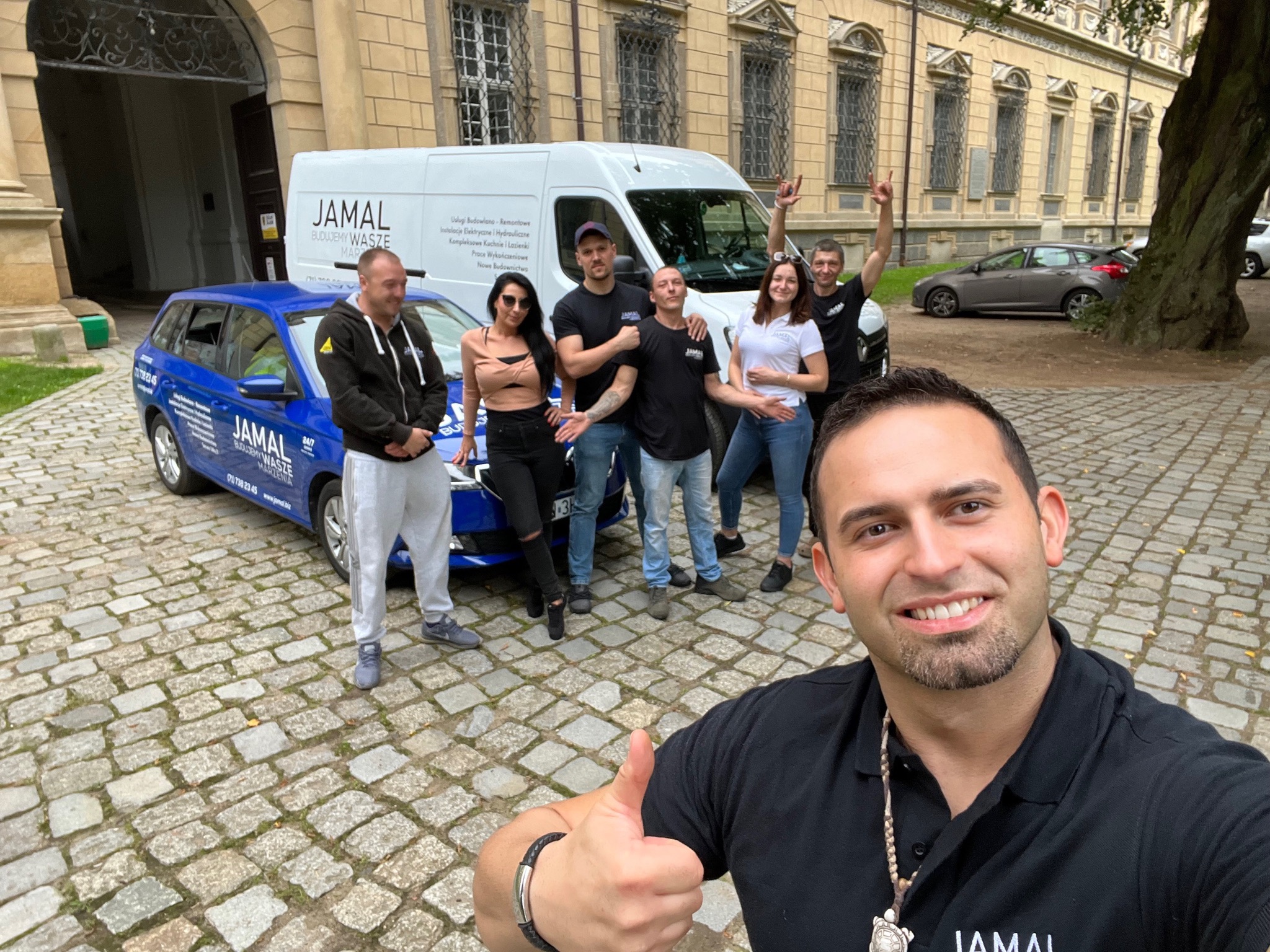Grupa pracowników firmy budowlanej Jamal pozuje do zdjęcia typu selfie na tle samochodów firmowych i budynku z cegły, jeden z mężczyzn pokazuje kciuk w górę.