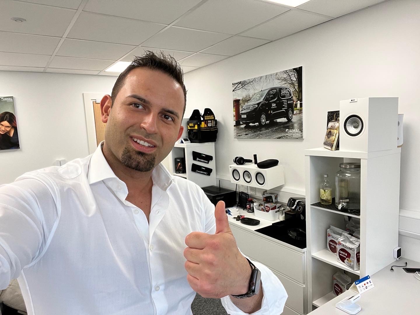 Uśmiechnięty mężczyzna w białej koszuli daje kciuk w górę na tle biura z półkami, na których znajdują się głośniki, słuchawki, ekspres do kawy i plakat z samochodem firmy Jamal Home.