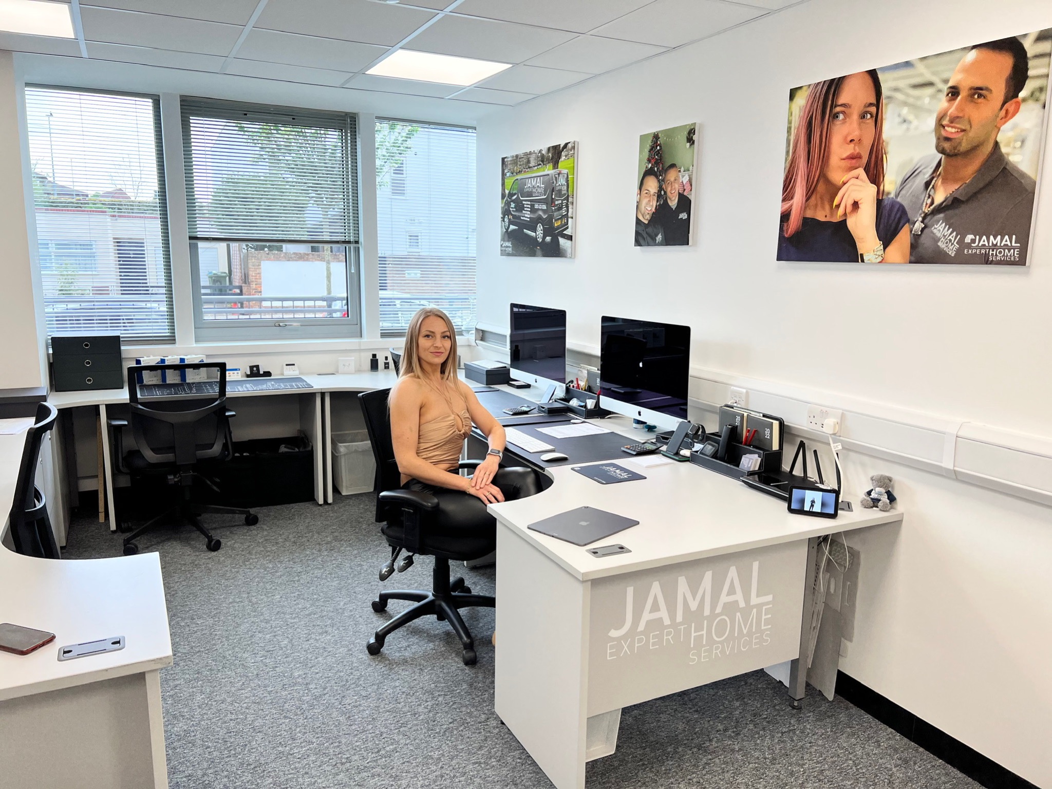 Wnętrze biura z blondynką siedzącą przy biurku z dwoma monitorami, na ścianach wiszą obrazy z logiem firmy Jamal Expert Home Services
