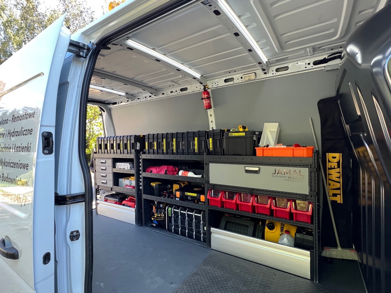 Wnętrze furgonetki serwisowej z profesjonalnym systemem regałów i pojemników na narzędzia, widoczny sprzęt marki DeWalt, gaśnica oraz logo firmy Jamal Budujemy Wasze Marzenia.