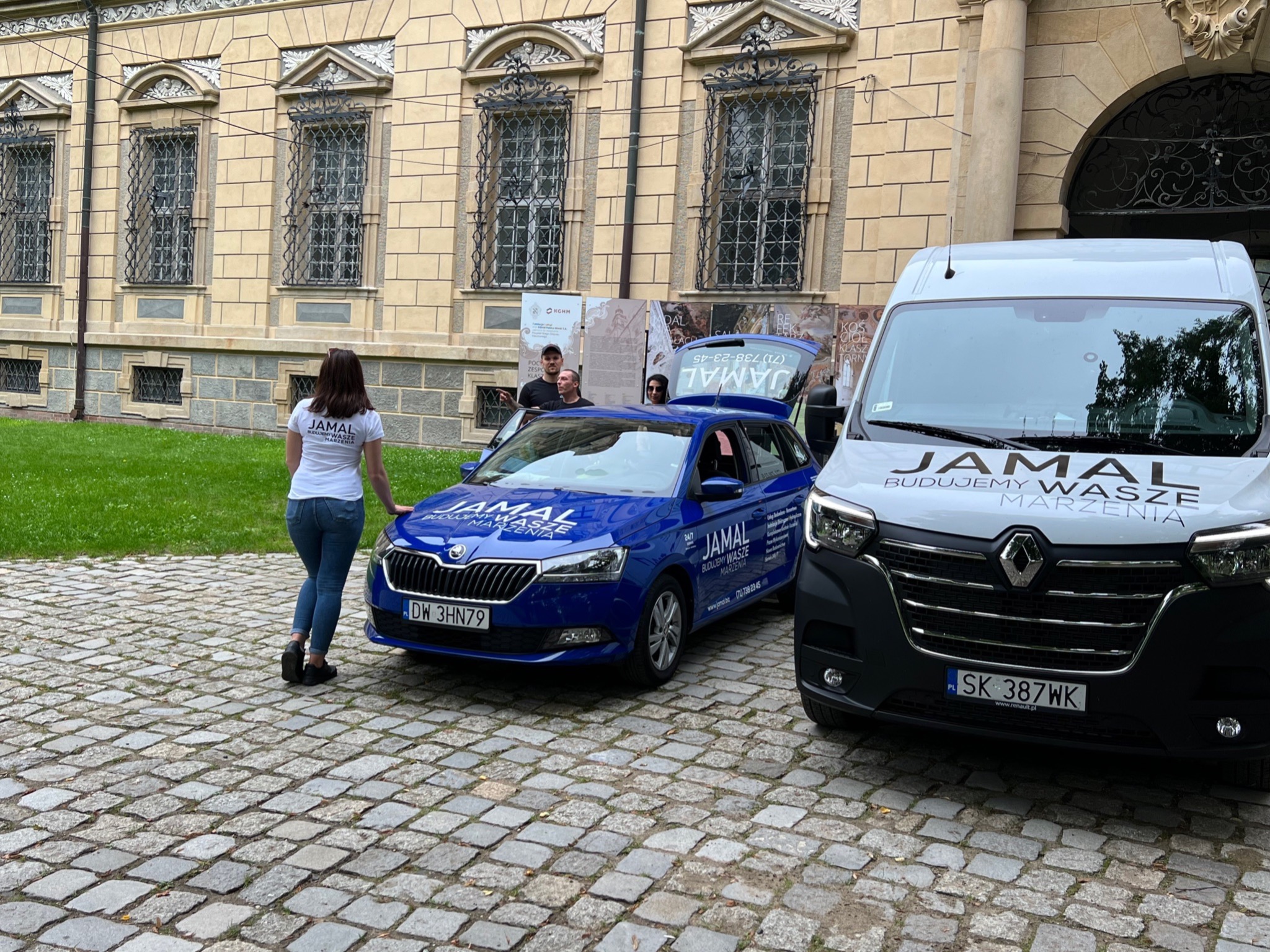 Dwa samochody firmy budowlanej Jamal zaparkowane na brukowanym dziedzińcu przed budynkiem z ozdobnymi oknami; na niebieskim samochodzie widoczne dane kontaktowe firmy.