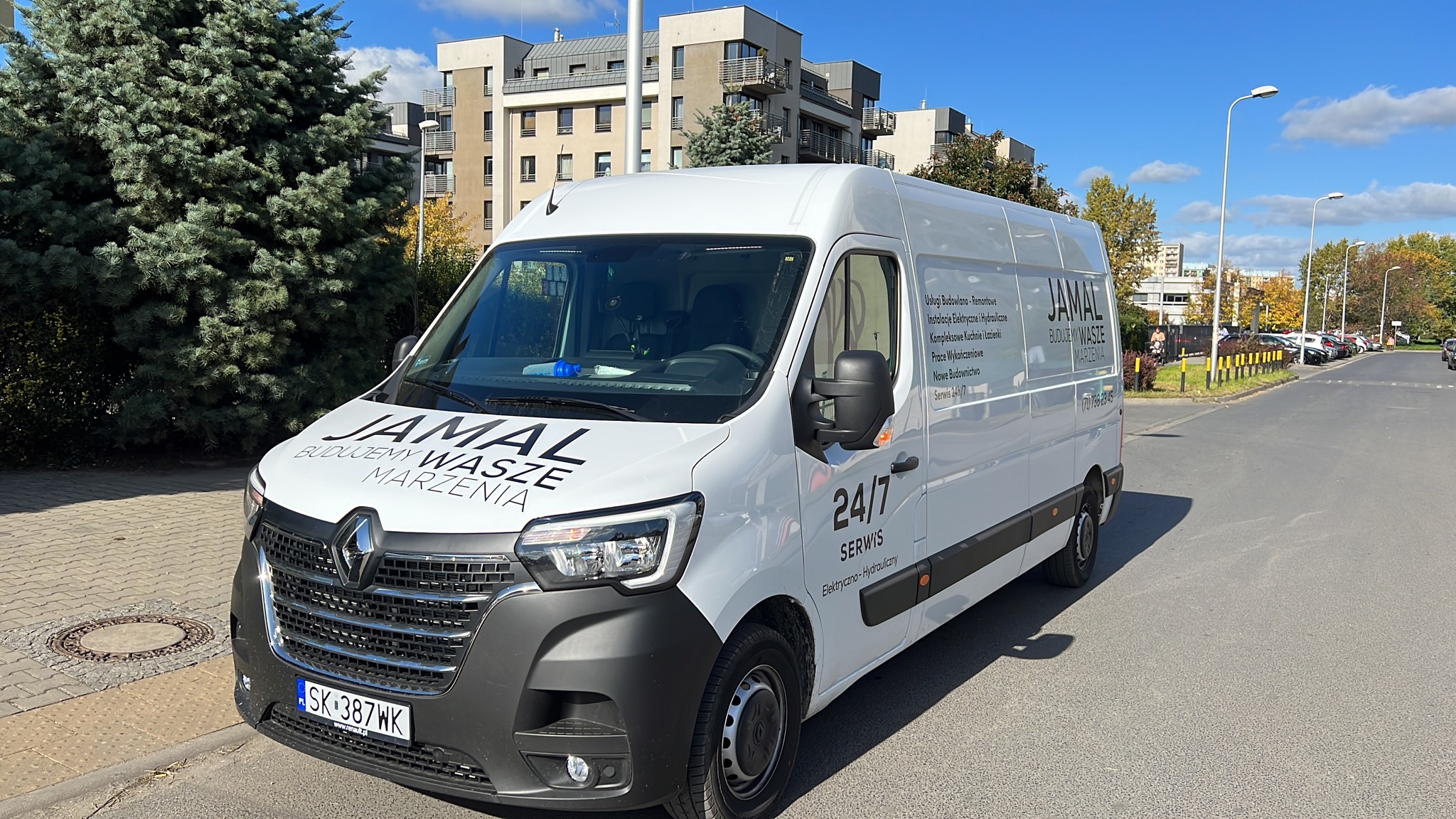 Biały samochód dostawczy firmy Jamal z napisem 'Budujemy Wasze Marzenia' i numerem telefonu, zaparkowany na ulicy w słoneczny dzień, na tle budynków mieszkalnych i drzew.
