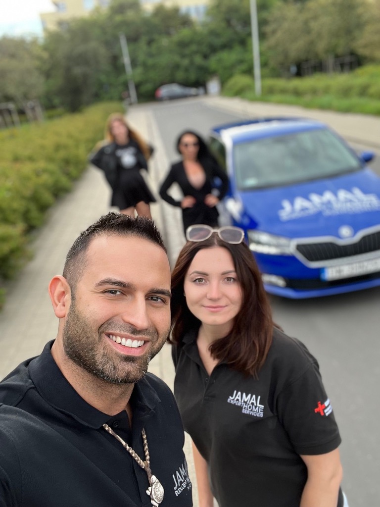 Selfie dwóch osób w czarnych koszulkach z logo firmy na tle niebieskiego samochodu z tym samym logo i trzech kobiet w tle.