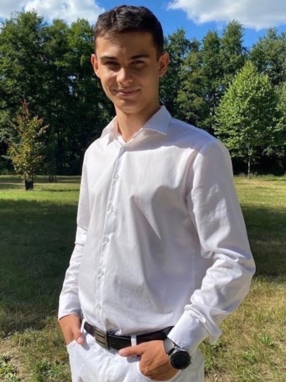 Młody mężczyzna w białej koszuli i spodniach, z zegarkiem na ręku, pozuje na tle zieleni w Tarnowie Podgórnym.