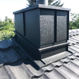 PlUAroof.pl - Szczegółowe ujęcie czarnej, metalowej obudowy komina na dachu z blachy falistej, z widocznymi elementami mocującymi i zielonym tłem drzew.