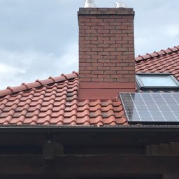 PlUAroof.pl - Czerwona dachówka ceramiczna na dachu z kominem z cegły, oknem dachowym i panelem solarnym w pochmurny dzień.