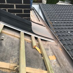 PlUAroof.pl - Montaż obróbek blacharskich przy kominie na dachu z dachówki ceramicznej; widoczne drewniane łaty, fragment czarnego komina z cegły i narzędzia dekarskie.