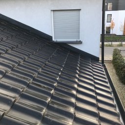 PlUAroof.pl - Błyszczące, czarne dachówki ceramiczne ułożone na dachu domu jednorodzinnego z oknem z roletą zewnętrzną i rynną wzdłuż krawędzi dachu.