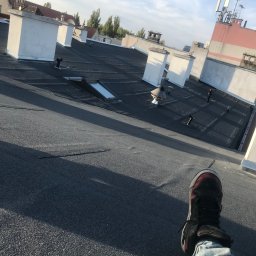 PlUAroof.pl - Widok z góry na czarny, płaski dach z kilkoma kominami i świetlikiem, w tle budynki miejskie, na pierwszym planie noga w jeansach i sportowym bucie.