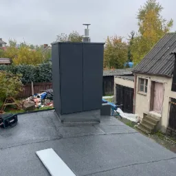 Dach pokryty papą termozgrzewalną z nowym, ciemnoszarym kominem z metalową czapką, widok z góry na tle zabudowań gospodarczych i jesiennego ogrodu.