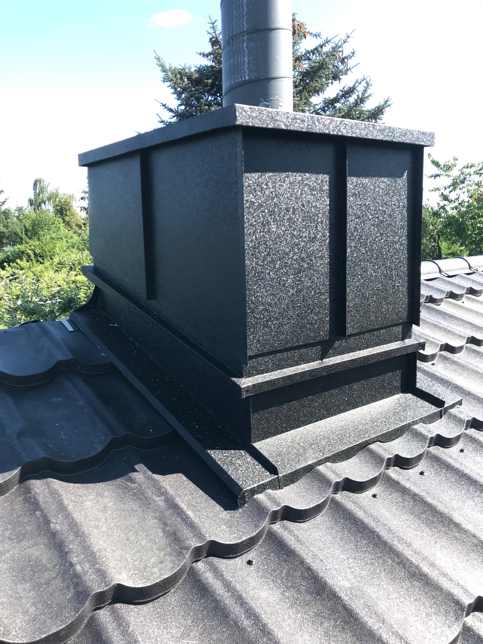 Szczegółowe ujęcie czarnej, metalowej obudowy komina na dachu z blachy falistej, z widocznymi elementami mocującymi i zielonym tłem drzew.