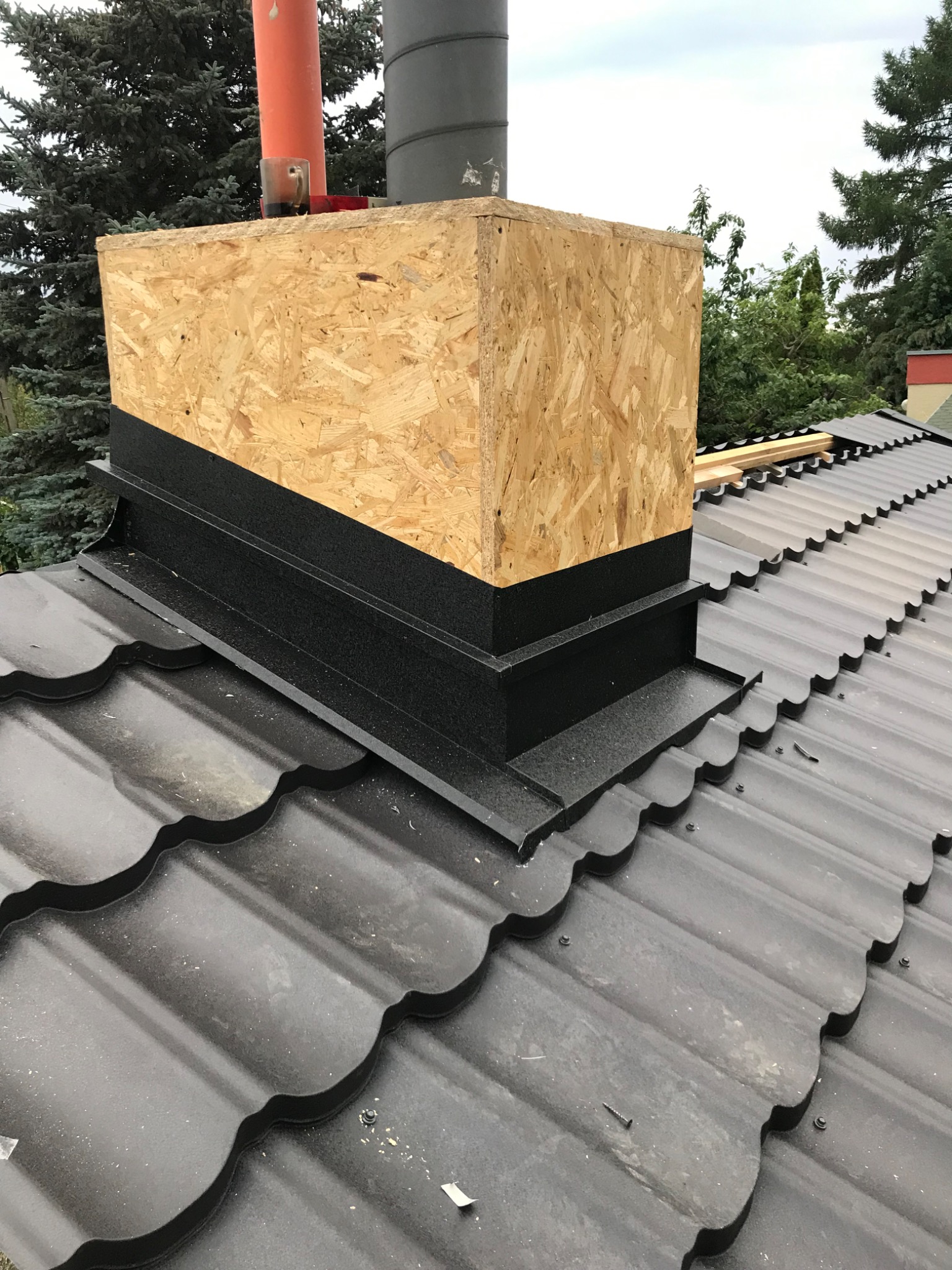 Obróbka komina z blachy i płyty OSB na dachu pokrytym metalową dachówką modułową w kolorze grafitowym.