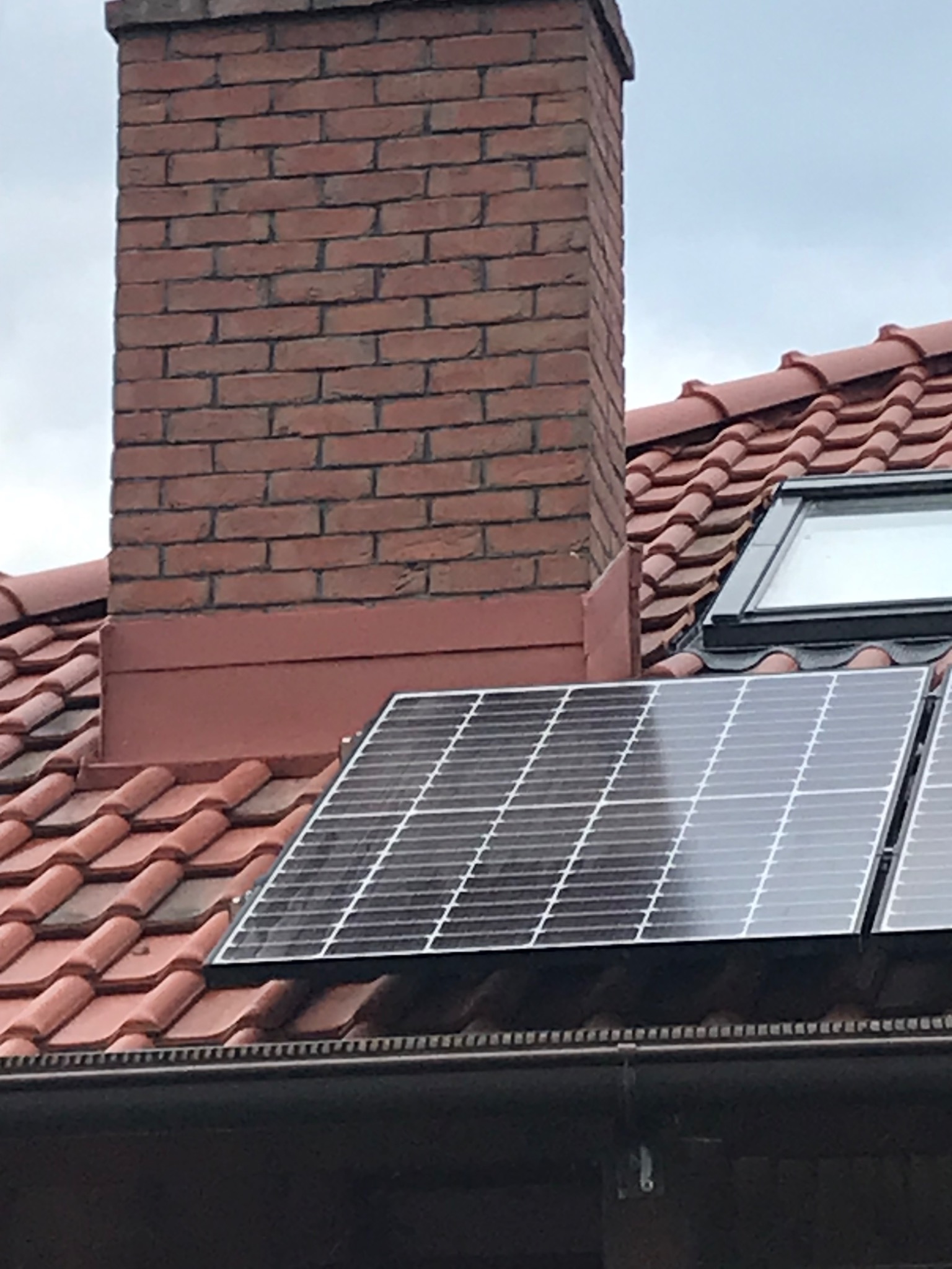 Czerwona dachówka ceramiczna z kominem z cegły, oknem dachowym i panelami słonecznymi w pochmurny dzień.