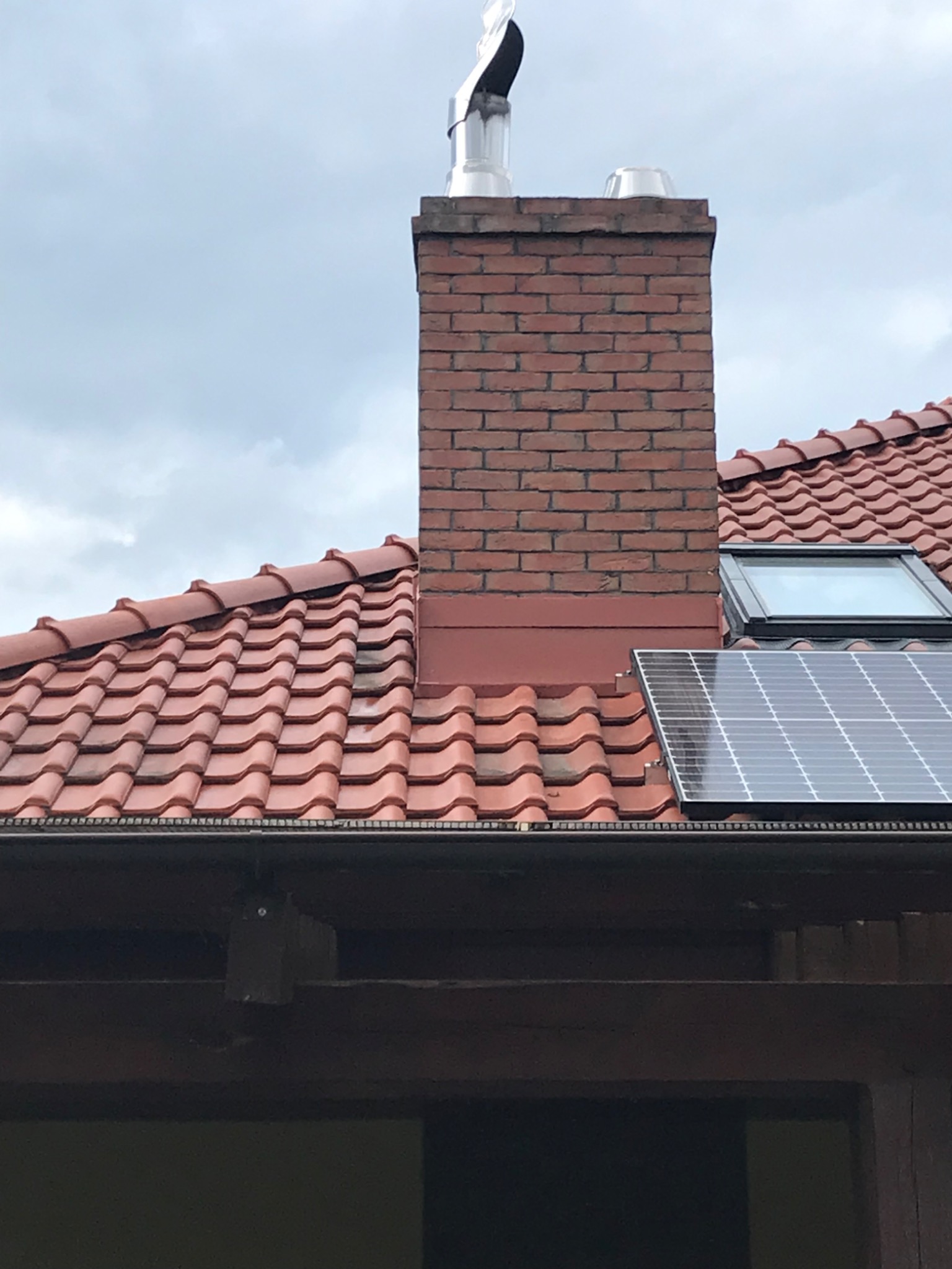 Czerwona dachówka ceramiczna na dachu z kominem z cegły, oknem dachowym i panelem solarnym w pochmurny dzień.
