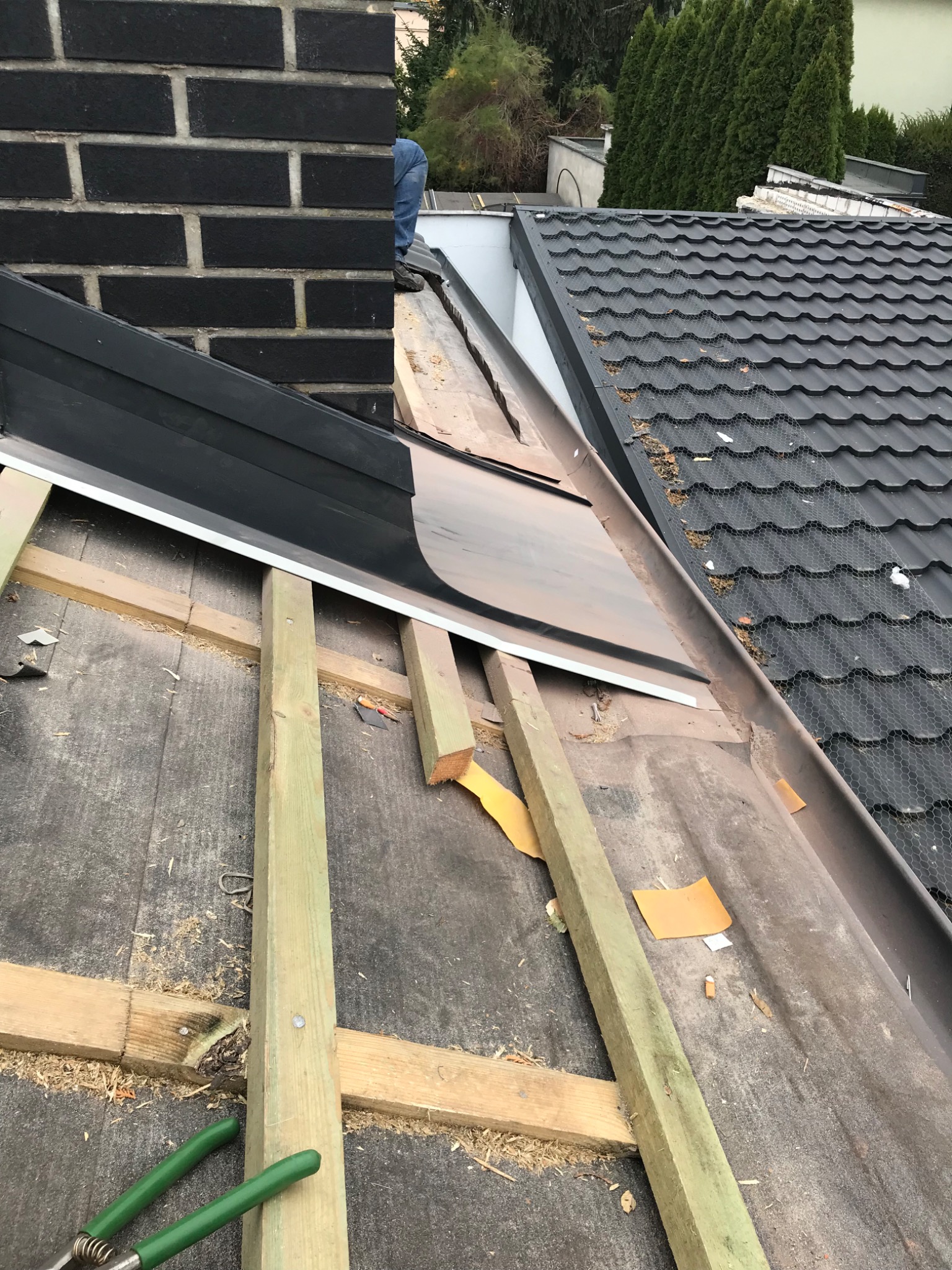 Montaż obróbek blacharskich przy kominie na dachu z dachówki ceramicznej; widoczne drewniane łaty, fragment czarnego komina z cegły i narzędzia dekarskie.