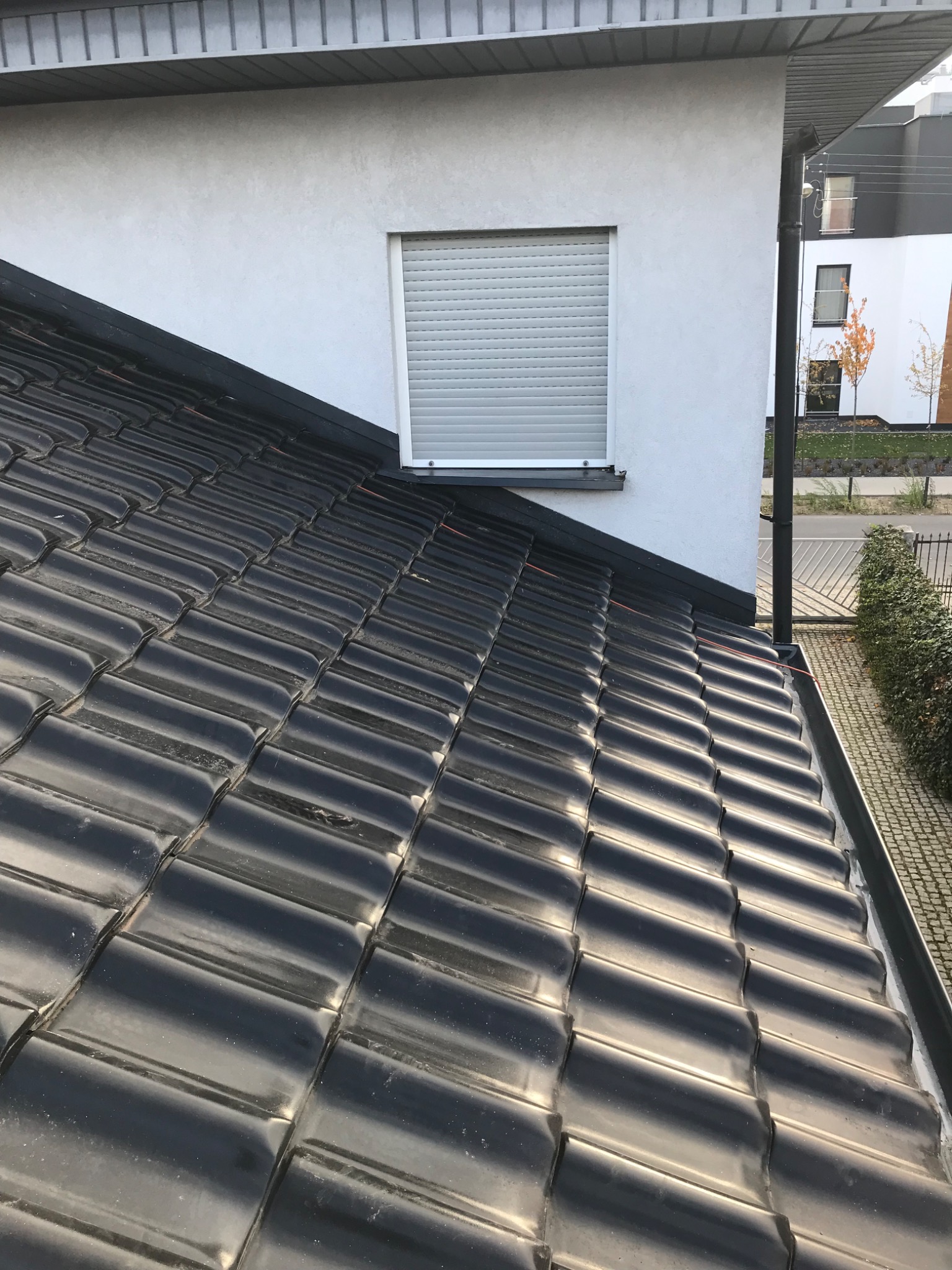 Błyszczące, czarne dachówki ceramiczne ułożone na dachu domu jednorodzinnego z oknem z roletą zewnętrzną i rynną wzdłuż krawędzi dachu.