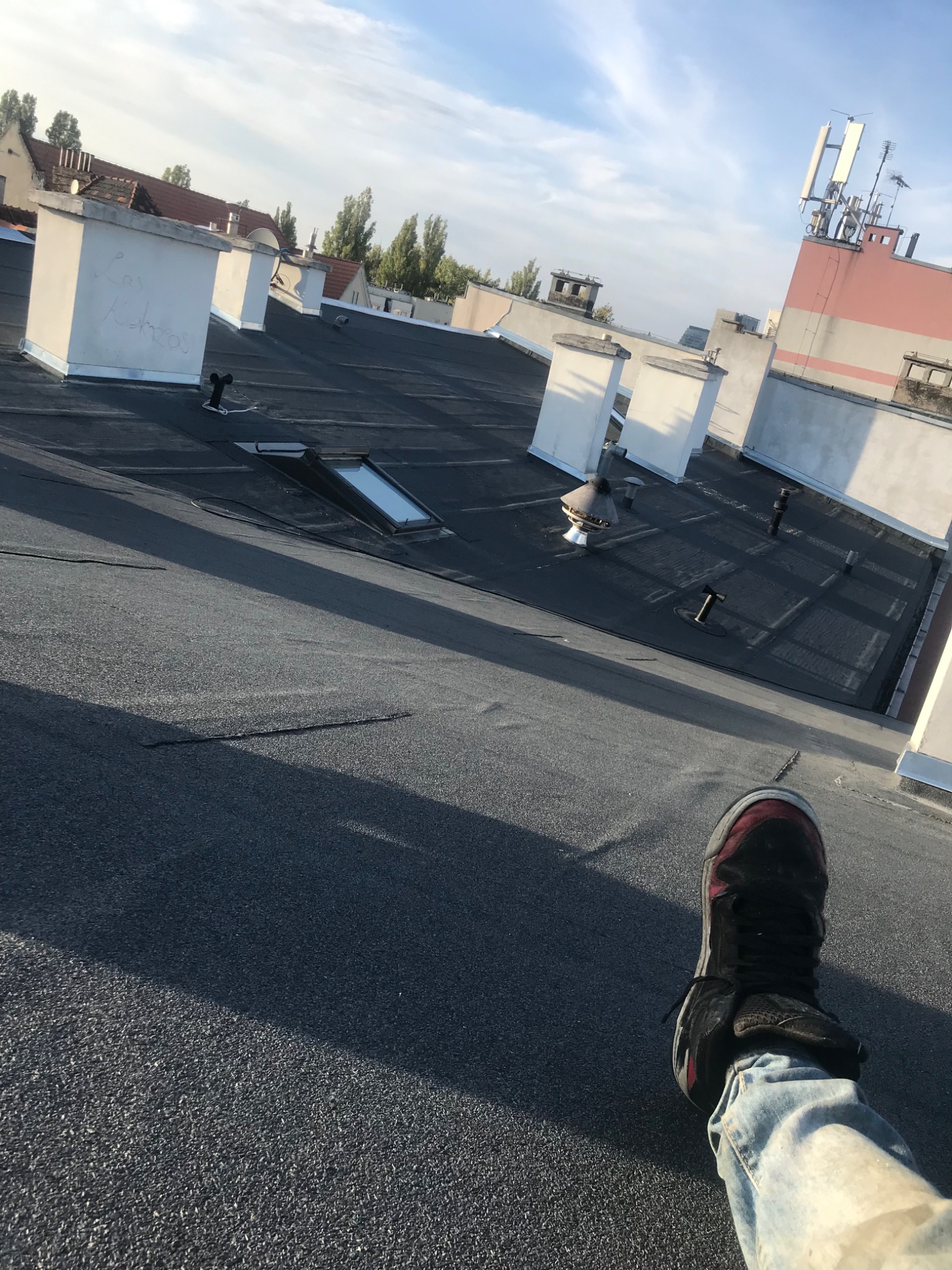Widok z góry na czarny, płaski dach z kilkoma kominami i świetlikiem, w tle budynki miejskie, na pierwszym planie noga w jeansach i sportowym bucie.