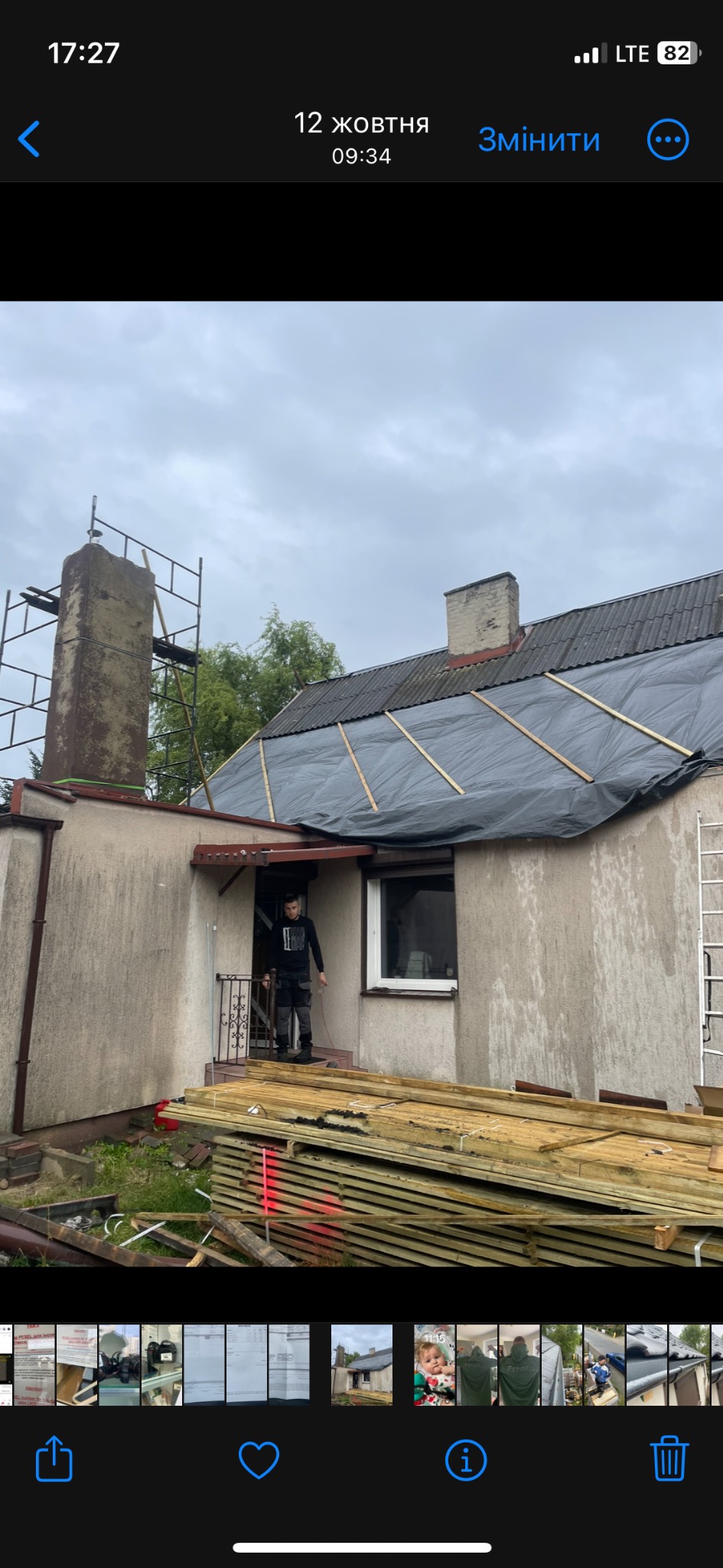 Dach zabezpieczony czarną folią przed deszczem, widoczne łaty drewniane, komin w rusztowaniu, stos drewnianych desek przed budynkiem, pracownik na ganku.