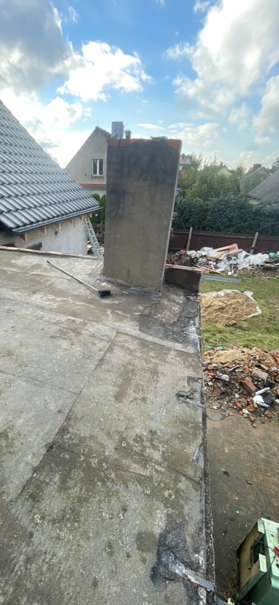 Widok z góry na płaski dach z kominem, częściowo zniszczony, z widocznymi śladami napraw. W tle dom z nowym dachem z dachówki i drabina oparta o ścianę. Na ziemi gruz i piasek.