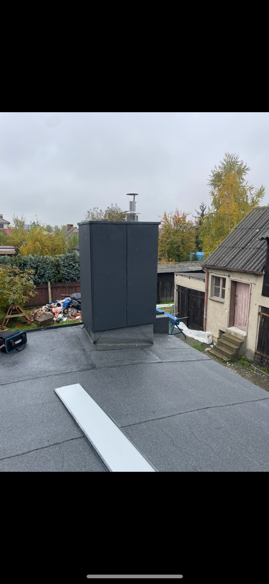 Dach pokryty papą termozgrzewalną z nowym, ciemnoszarym kominem z metalową czapką, widok z góry na tle zabudowań gospodarczych i jesiennego ogrodu.