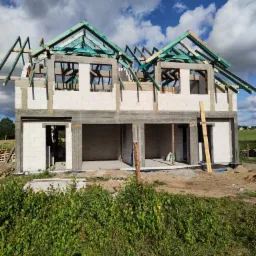 Budowa bliźniaka w stanie surowym otwartym: ściany z betonu komórkowego, zielona więźba dachowa, widoczne otwory okienne i drzwiowe, na pierwszym planie zarośla.