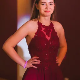 Portret młodej kobiety w bordowej, zdobionej sukni wieczorowej z koronkową górą, pozującej z rękami na biodrach, z bransoletką świecącą na nadgarstku.