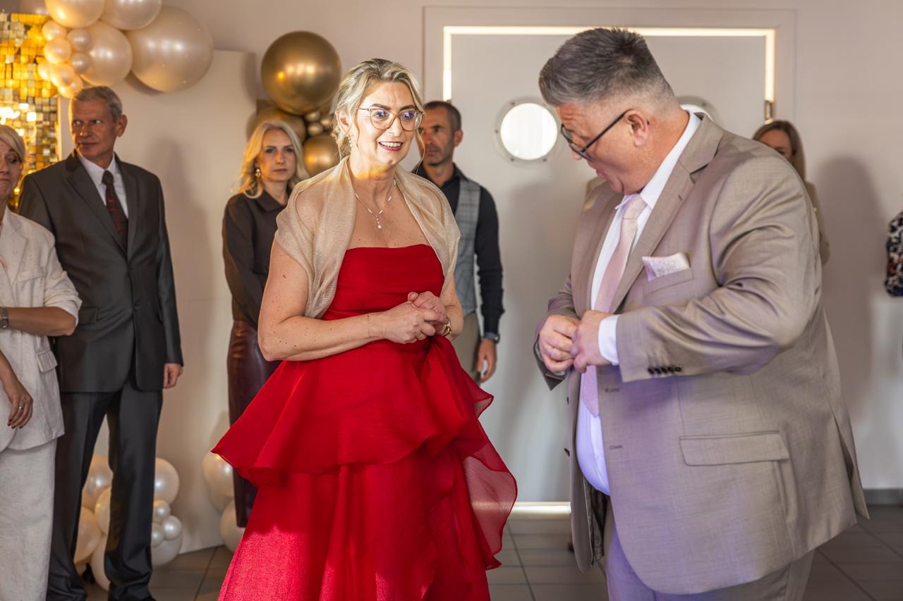 Kobieta w czerwonej sukni na tle dekoracji balonowych, w otoczeniu gości. Mężczyzna w garniturze zapina guzik. Ujęcie z imprezy okolicznościowej.