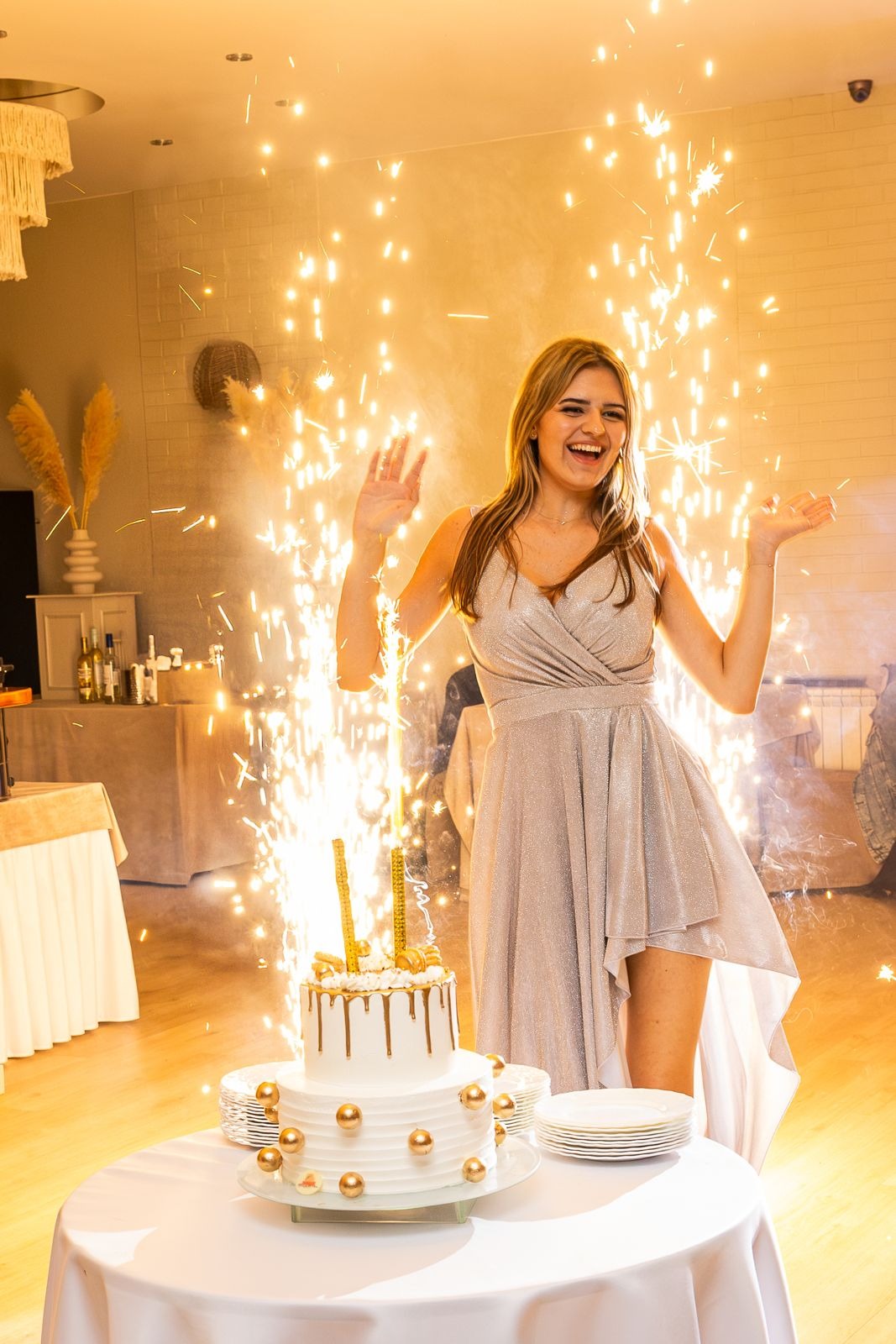 Uśmiechnięta kobieta w srebrnej sukience pozuje przy stole z tortem urodzinowym, otoczona fontanną iskier. Jasne oświetlenie, radosna atmosfera.
