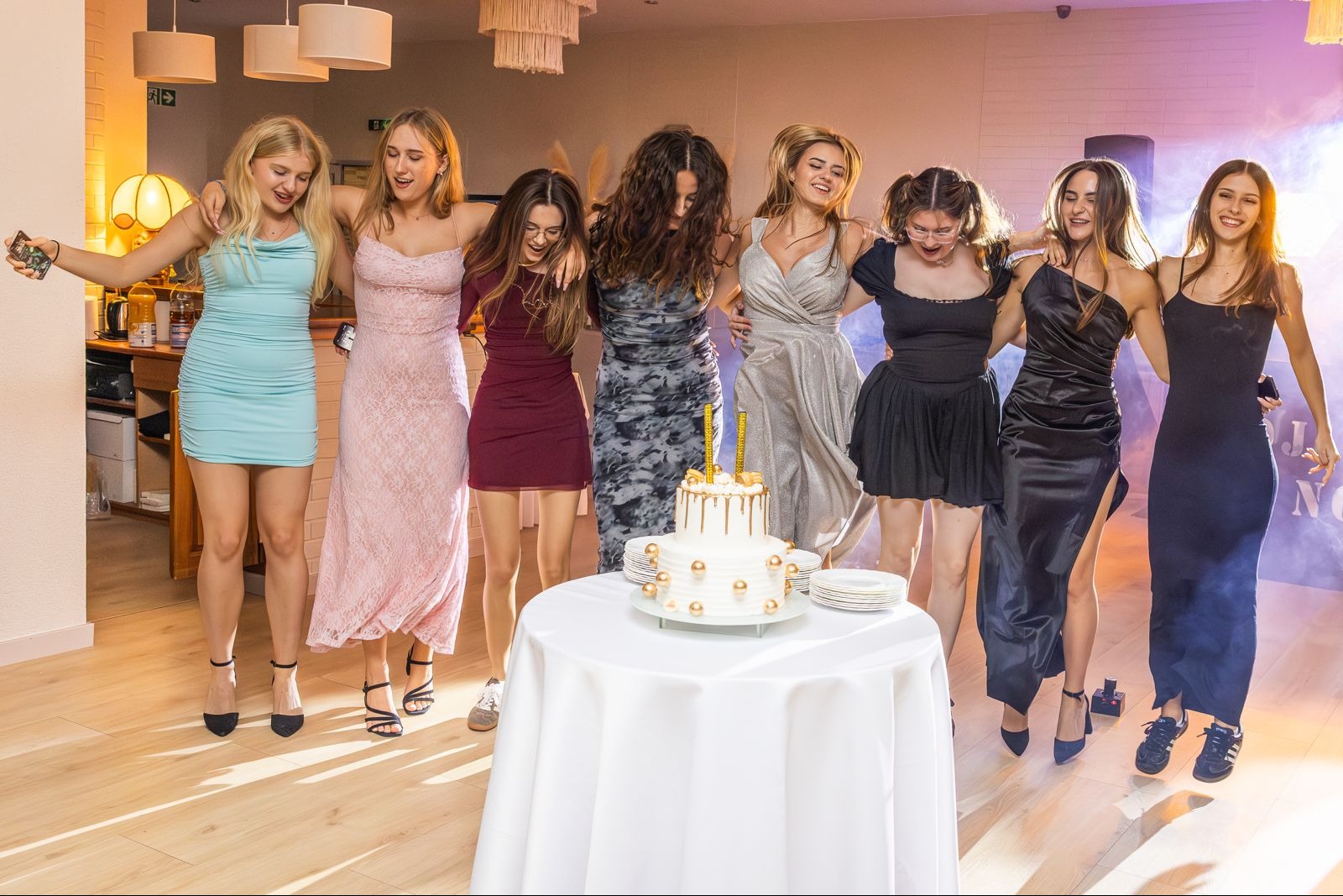 Grupa ośmiu młodych kobiet w sukienkach, stojących wokół stołu z tortem urodzinowym, w jasnym wnętrzu z dekoracjami i subtelnym oświetleniem.