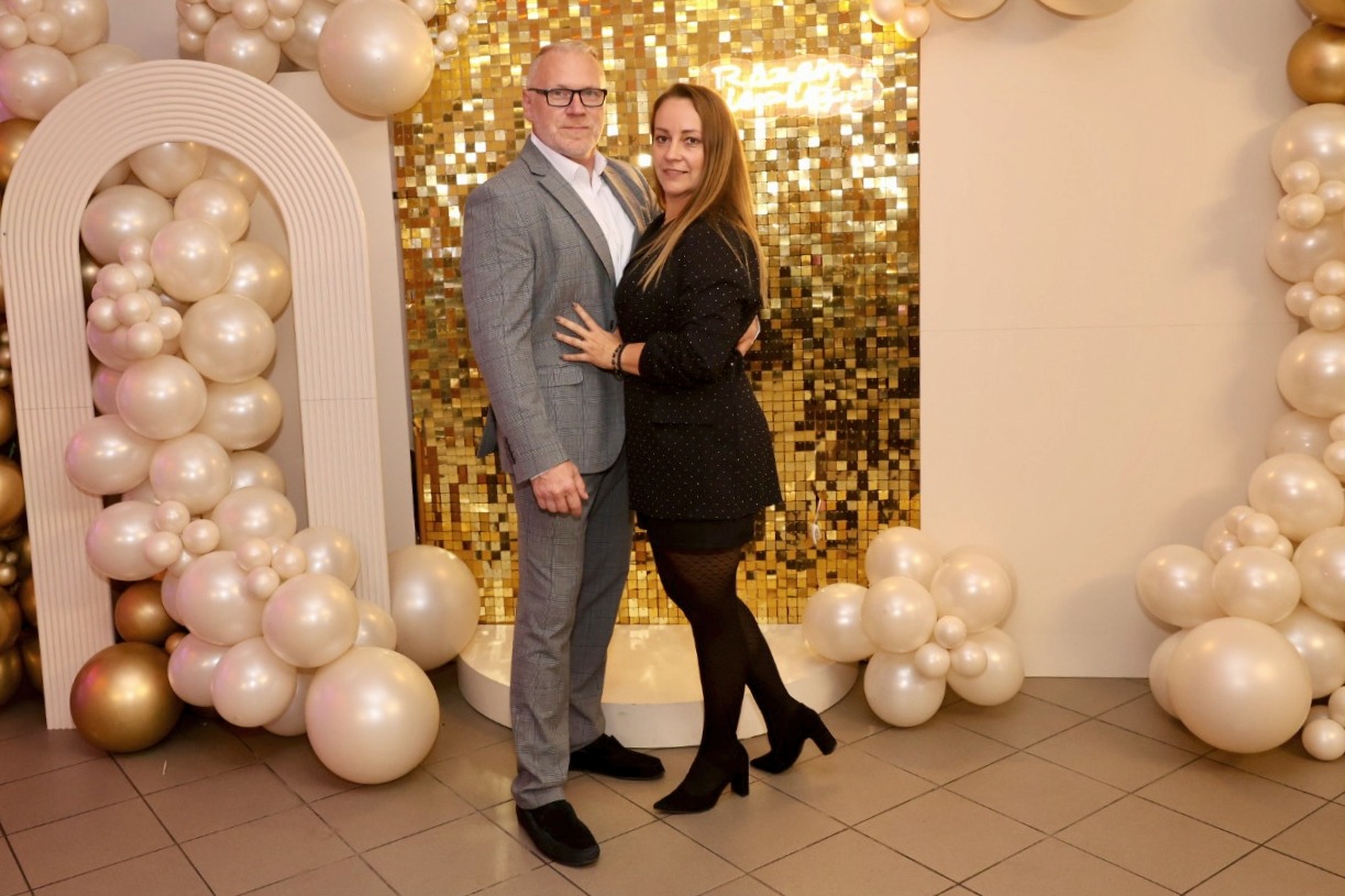 Para pozuje na tle złotej ściany z cekinów i dekoracji z białych balonów. Mężczyzna w szarej marynarce, kobieta w czarnej sukience. Uśmiechnięci, eleganccy.