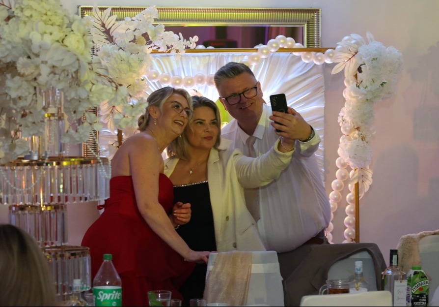 Trzy osoby robią selfie na tle dekoracji ślubnej z białych kwiatów i pereł. Kobieta w czerwonej sukience, kobieta w czarnej sukience i mężczyzna w białej koszuli.