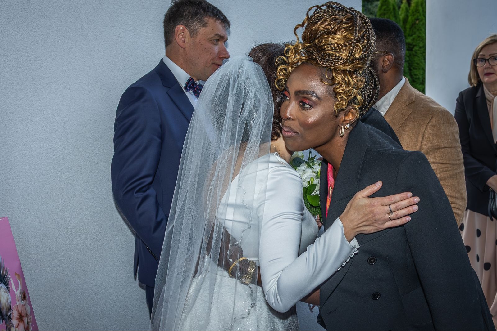 Panna młoda w białej sukni i welonie otrzymuje gratulacje od kobiety w czarnym płaszczu. W tle goście weselni w eleganckich strojach.