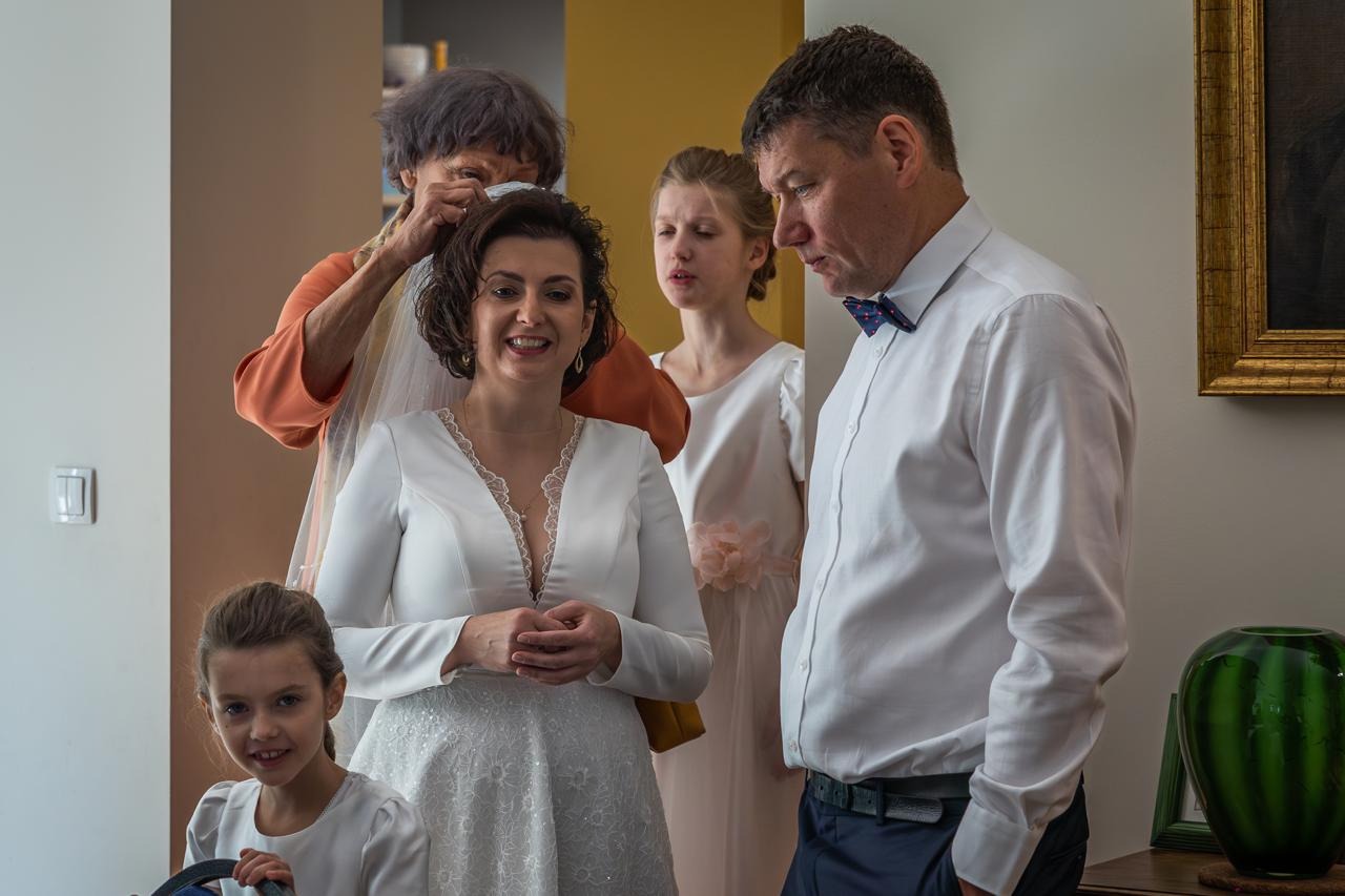 Panna młoda w białej sukni, poprawiana przez starszą kobietę, obok ojciec i dziewczynki w jasnych sukienkach. Przygotowania do ślubu w jasnym wnętrzu.