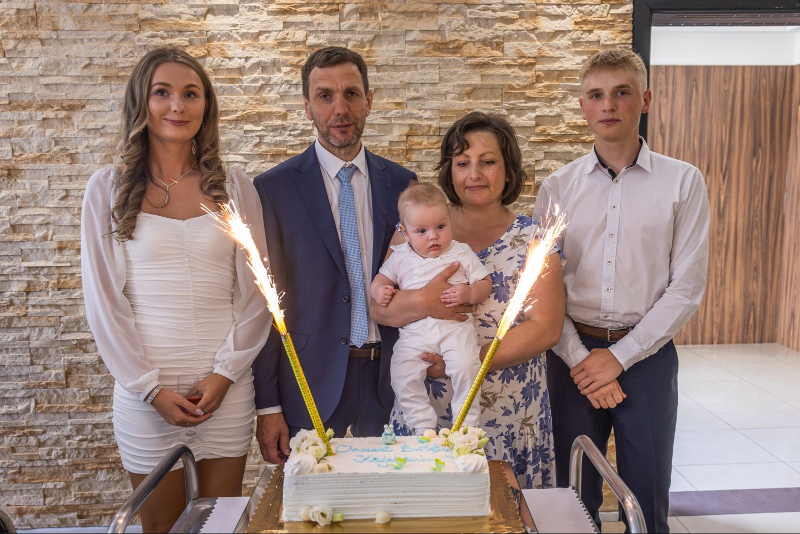 Rodzinne zdjęcie z uroczystości chrztu, tort ze świecami fontannowymi, rodzice i dzieci w eleganckich strojach na tle ściany z kamienia dekoracyjnego.