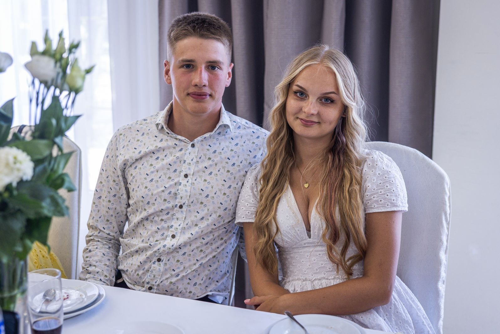 Para młoda siedzi przy stole podczas przyjęcia weselnego w Brzezinach. Panna młoda w białej sukience, pan młody w koszuli we wzory. Dekoracje kwiatowe na stole.