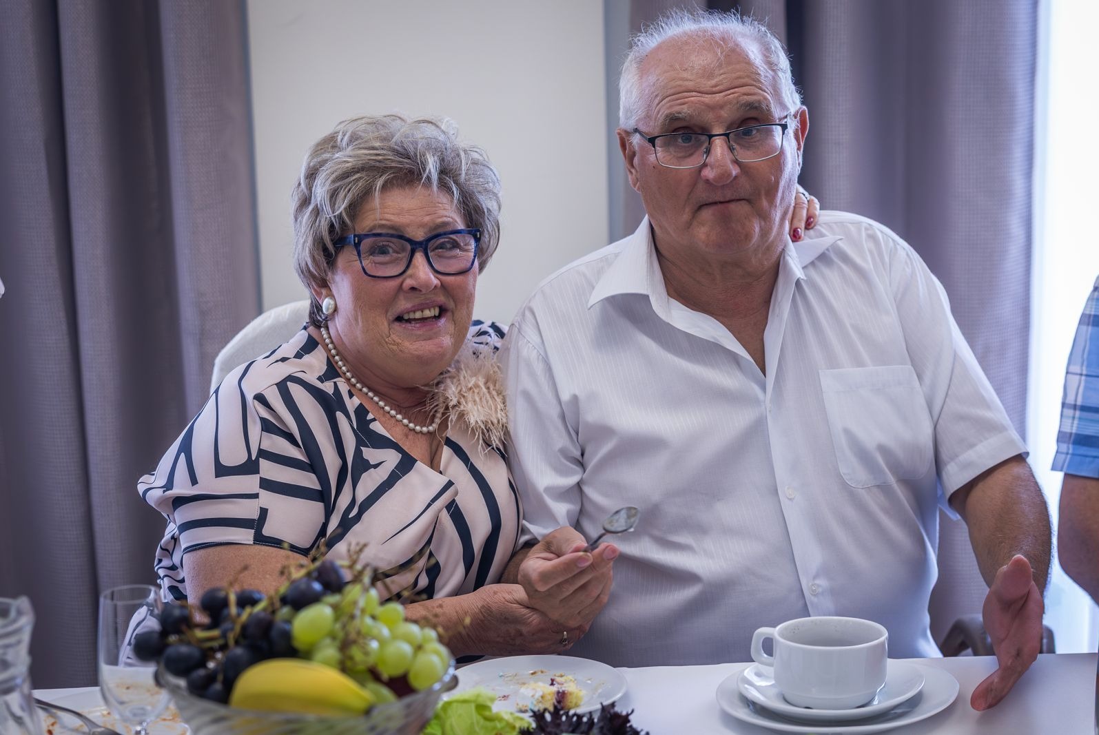 Uśmiechnięta para seniorów siedzi przy stole z owocami i kawą. Kobieta w okularach i perłach, mężczyzna w białej koszuli. Zdjęcie z uroczystości rodzinnej.