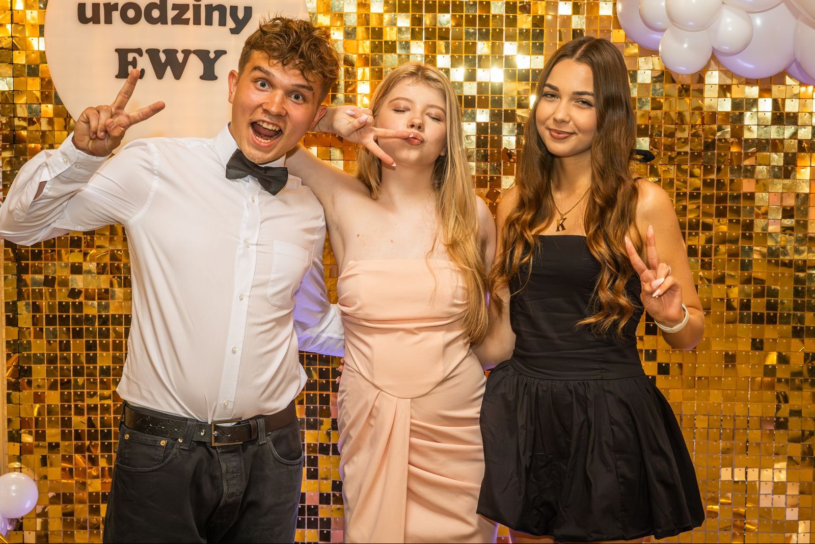 Radosne trio pozuje na tle złotej ściany z cekinami podczas urodzin. Mężczyzna w muszce i dwie kobiety w sukienkach prezentują gest zwycięstwa.
