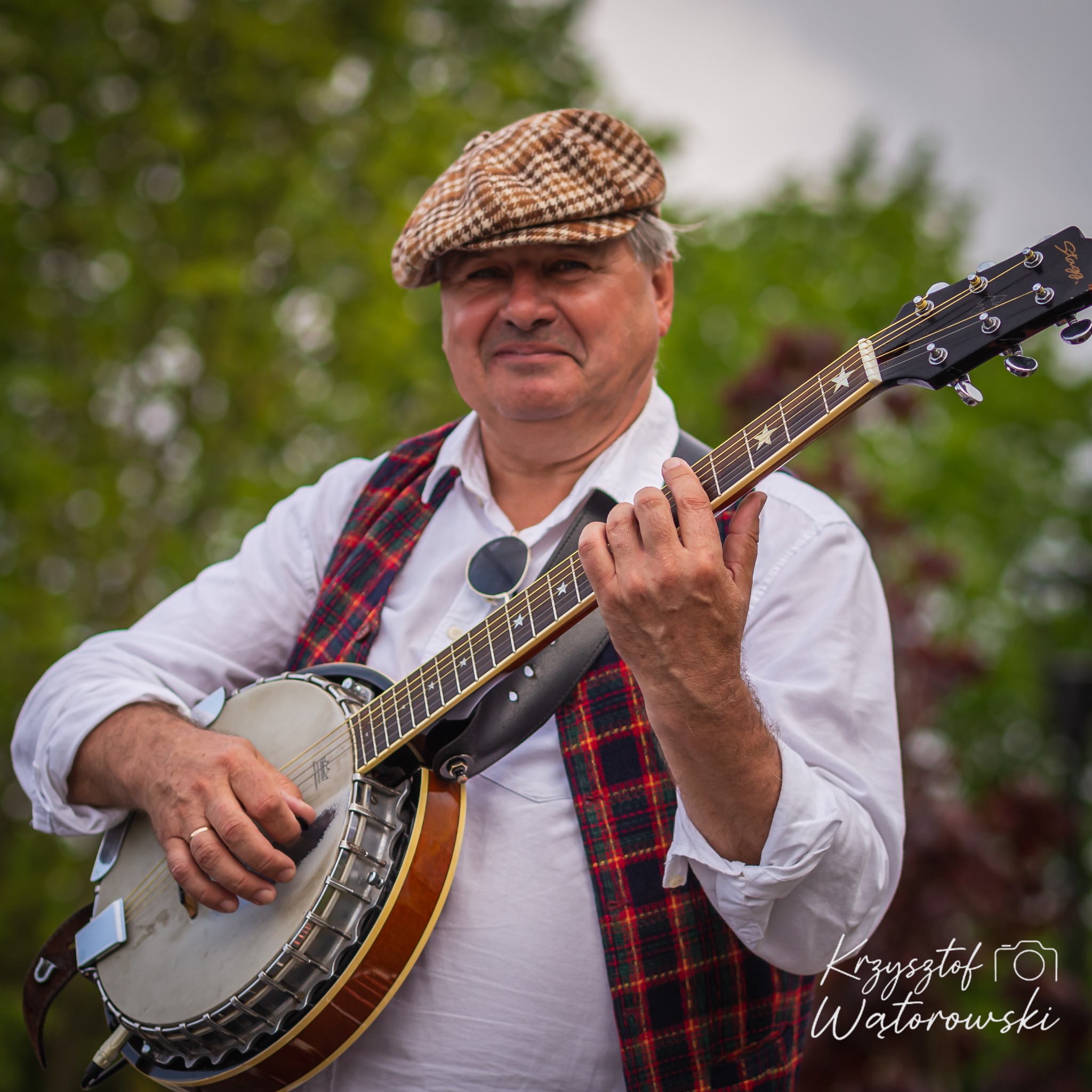 Uśmiechnięty muzyk w kaszkiecie gra na banjo, ubrany w białą koszulę i kraciastą kamizelkę. Tło rozmyte, zielone. Widoczny podpis fotografa w prawym dolnym rogu.