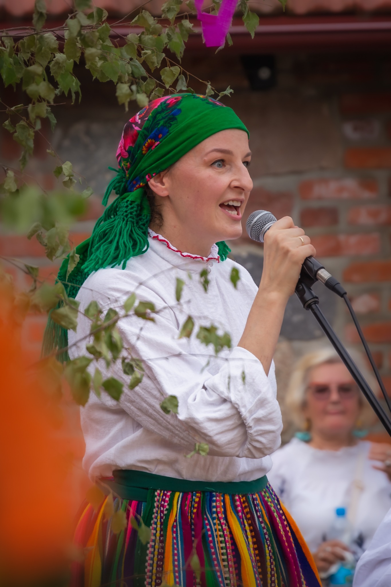Kobieta w ludowym stroju śpiewa do mikrofonu na tle ceglanej ściany i zieleni. Uśmiech, kolorowa chusta i spódnica, mikrofon w dłoni. Część twarzy zasłaniają liście.