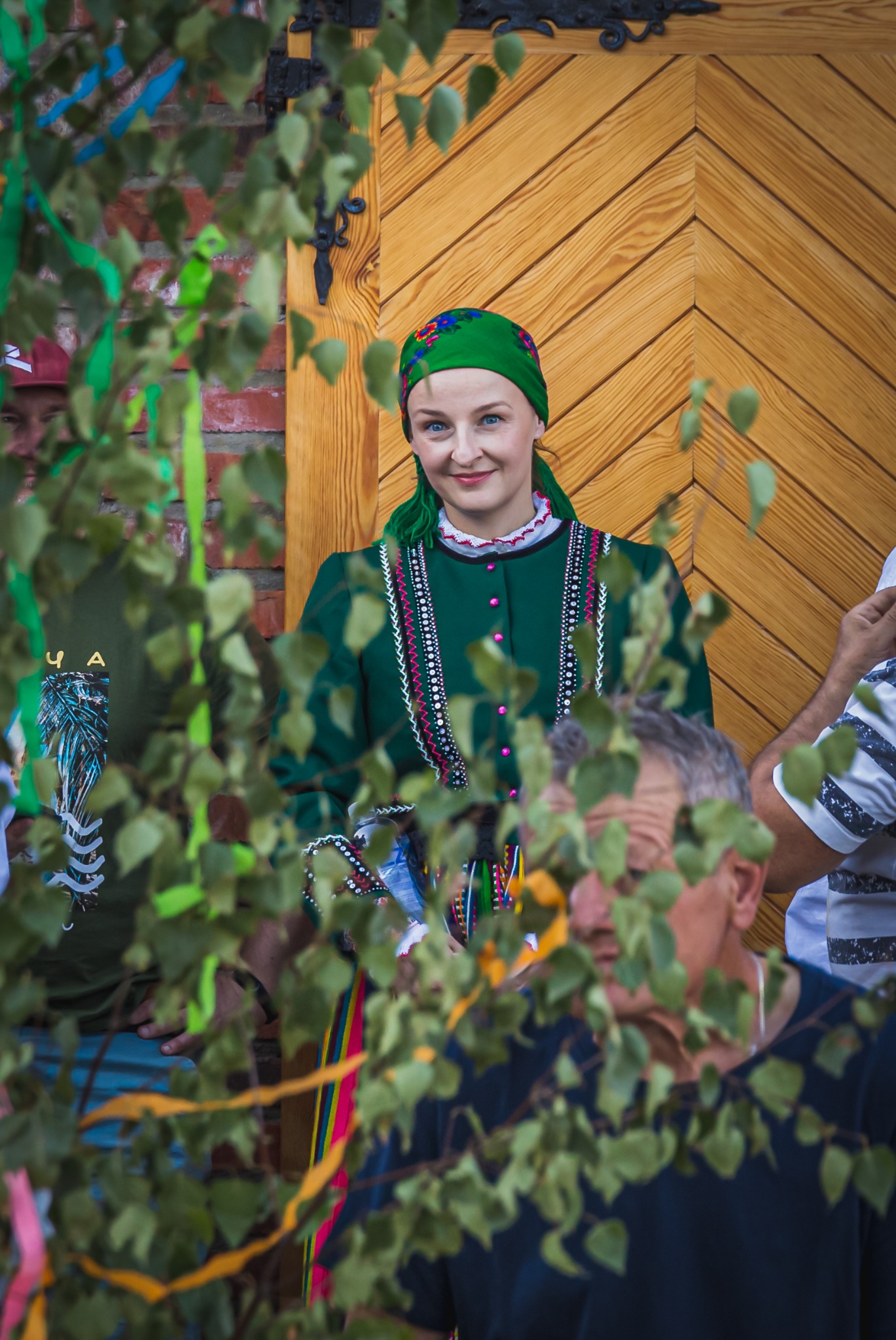 Uśmiechnięta kobieta w zielonym stroju ludowym i chustce na głowie, pozuje na tle drewnianych drzwi, częściowo zasłonięta gałęziami drzewa.
