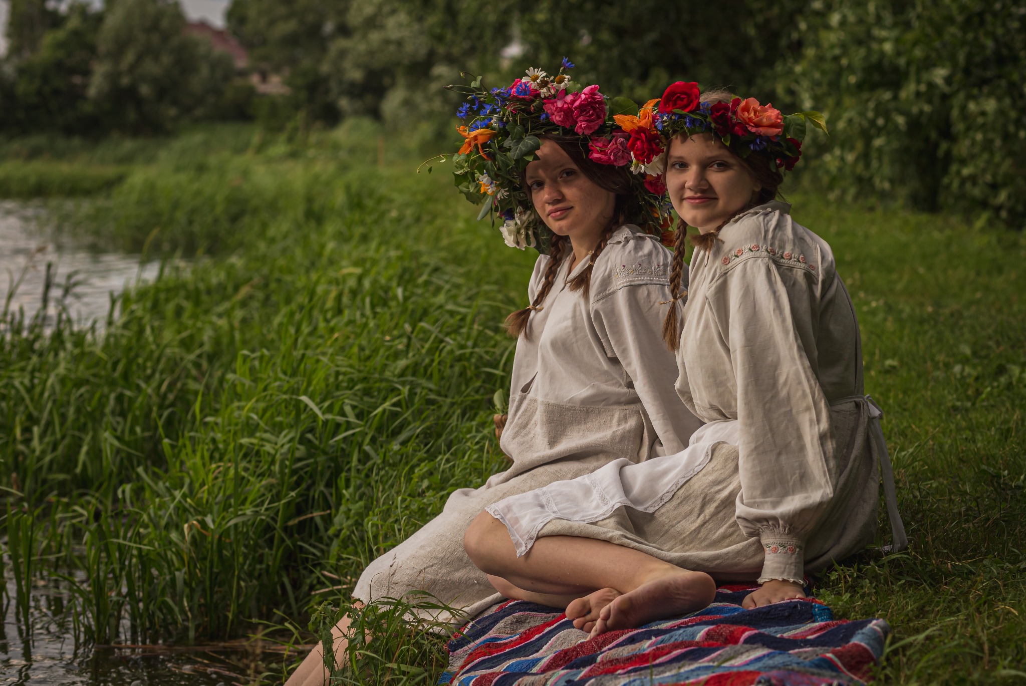 Dwie dziewczyny w ludowych strojach i wiankach siedzą na kocu nad brzegiem rzeki, otoczone zielenią. Ujęcie z bliska, naturalne światło, spokojna atmosfera.