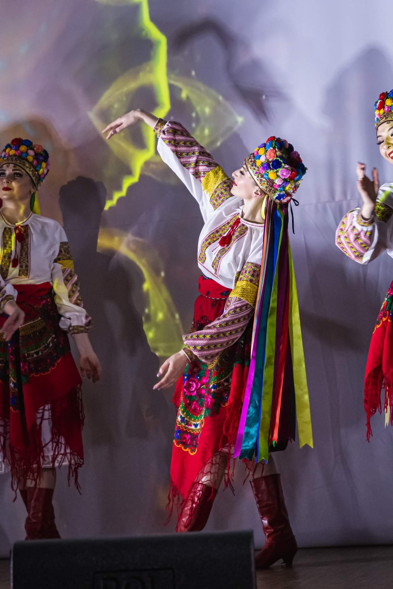 Zespół w tradycyjnych, ukraińskich strojach ludowych prezentuje układ taneczny na scenie z abstrakcyjnym tłem. Kolorowe wstążki i hafty zdobią stroje.